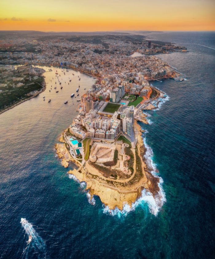 Aerial view of a coastal city at sunset, highlighting a peninsula surrounded by blue waters, dotted with boats. The cityscape features a mix of modern buildings and green spaces, with rocky shores and waves crashing near the land.