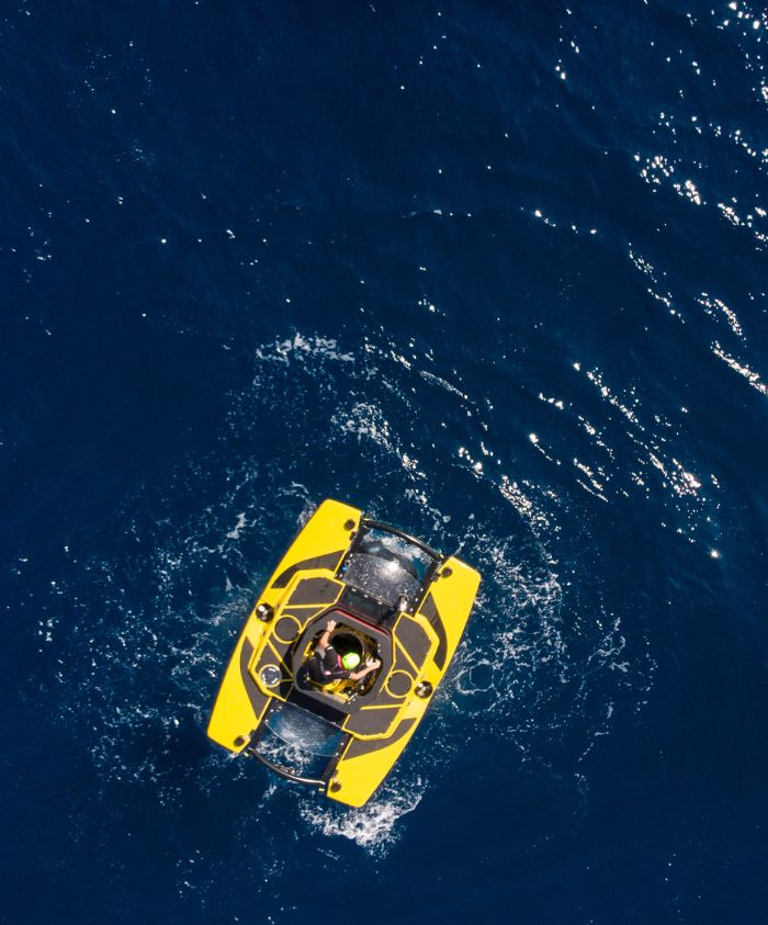 A bright yellow speedboat navigates through deep blue water, creating ripples around it. Two people are seated inside the boat, enjoying their time on the water.
