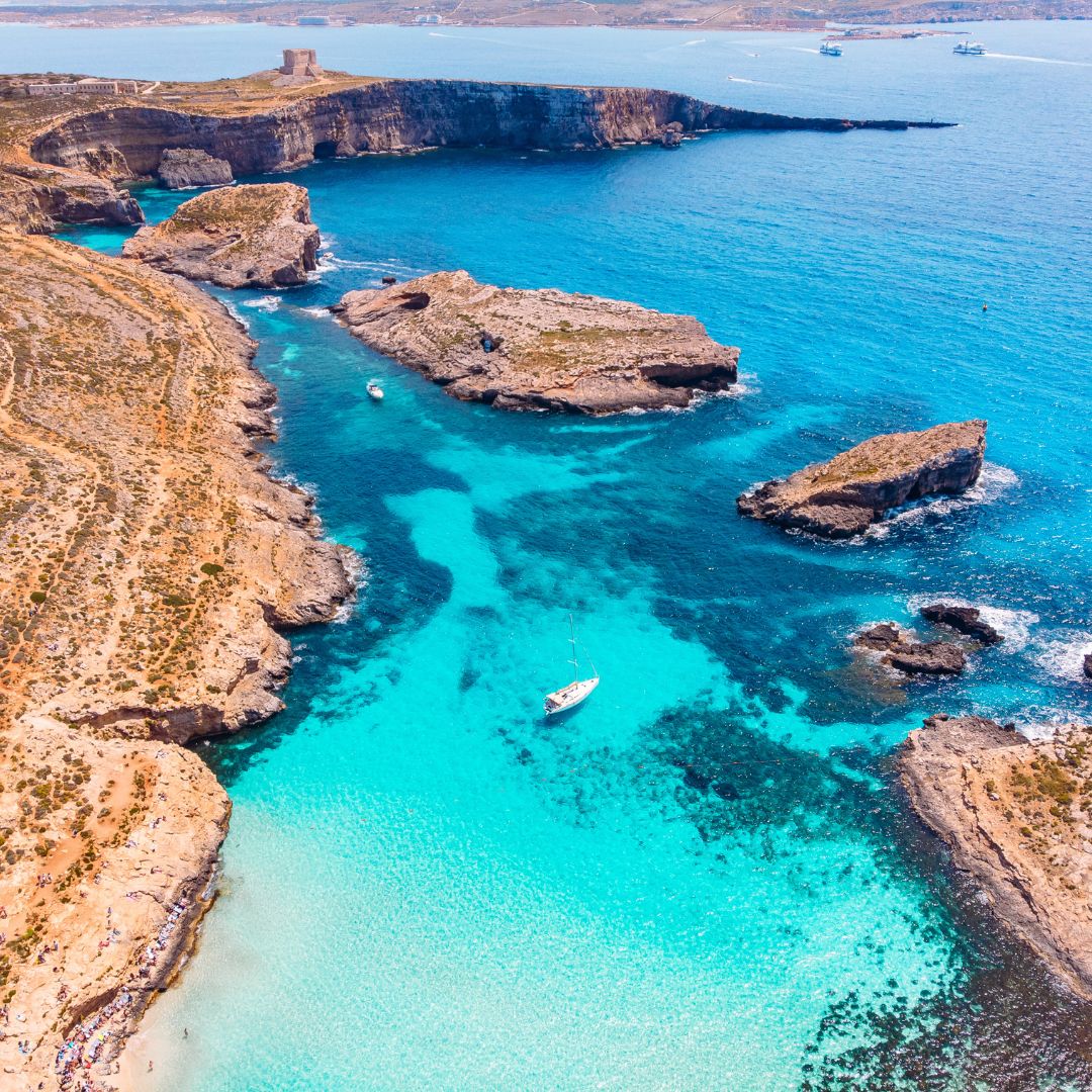 A serene coastline with rocky formations and clear turquoise waters. A single sailboat is anchored near the shore, surrounded by small islands and gentle waves. In the distance, a tower overlooks the landscape.