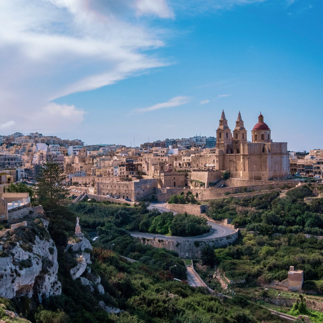 A scenic view of a hillside town with historic buildings, including a church with twin towers and a red dome, surrounded by greenery and a winding road. Clear blue sky overhead.