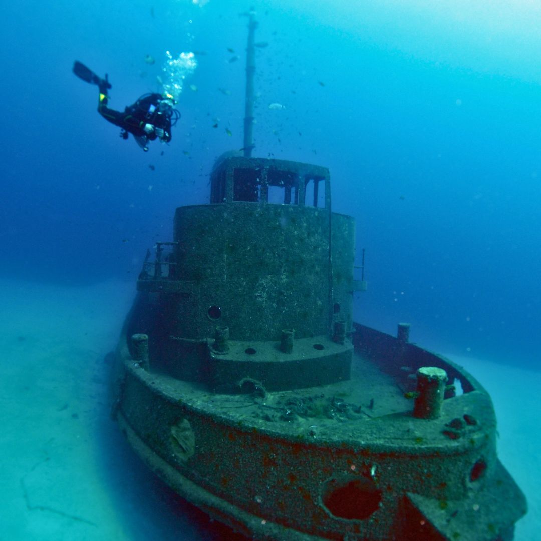 A diver explores a sunken tugboat covered in marine life and coral, partially submerged in clear blue water. Bubbles rise around the diver as they navigate the underwater scene.