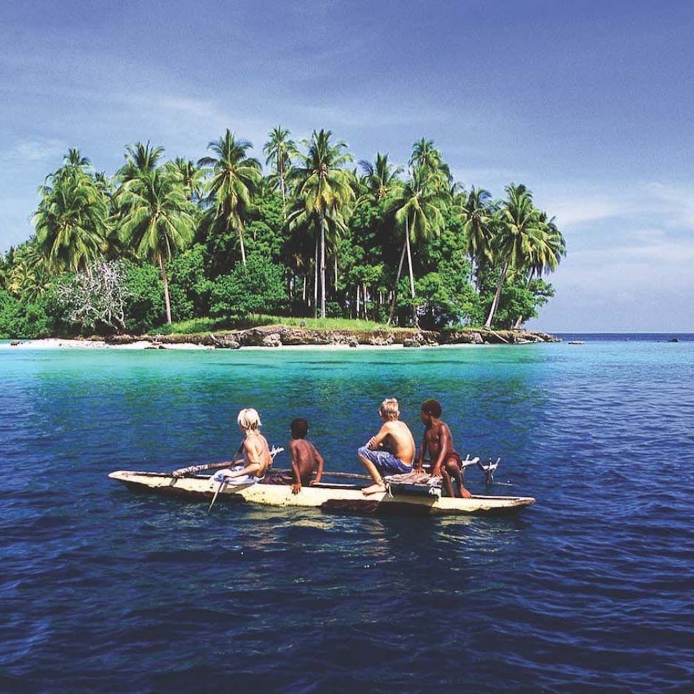 A group of four boys paddle a small canoe in clear turquoise waters near a lush, palm-fringed island under a bright blue sky.