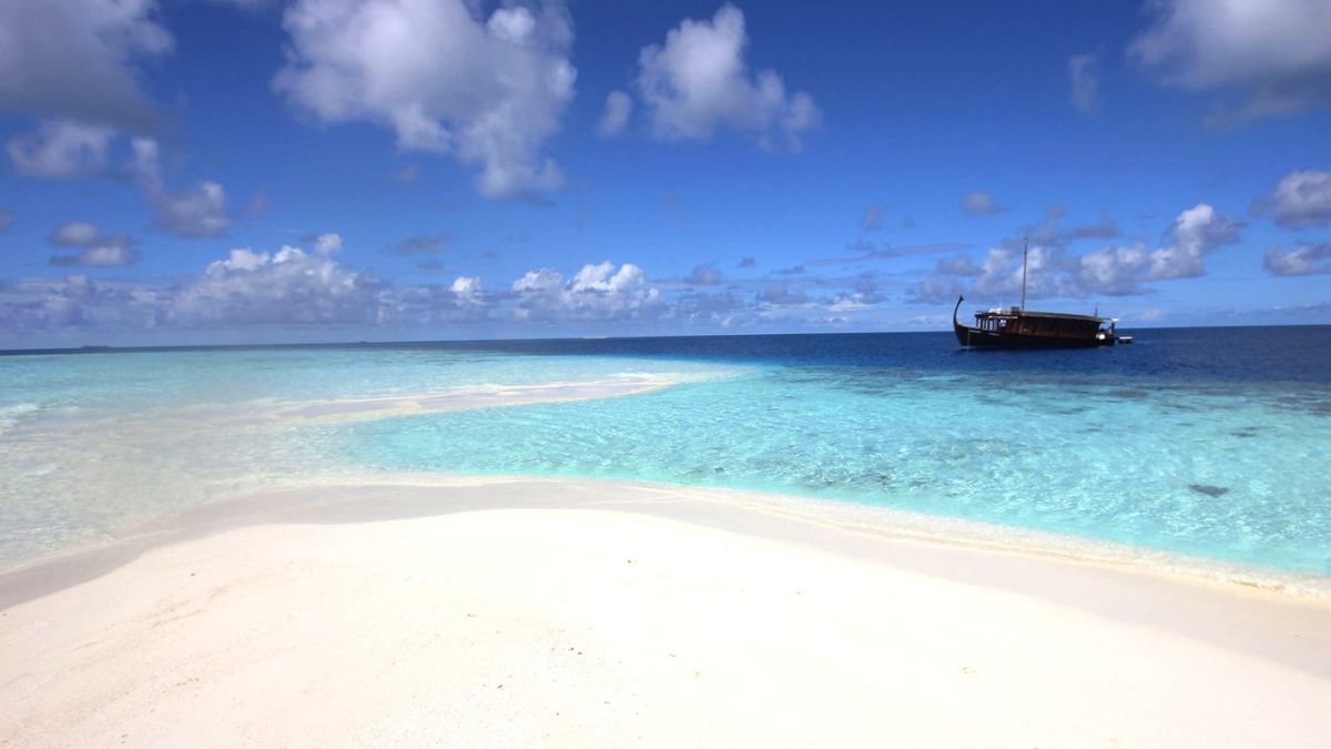 A serene beach scene with soft white sand and clear turquoise waters. A wooden boat is anchored in the distance against a backdrop of puffy clouds and a vibrant blue sky.