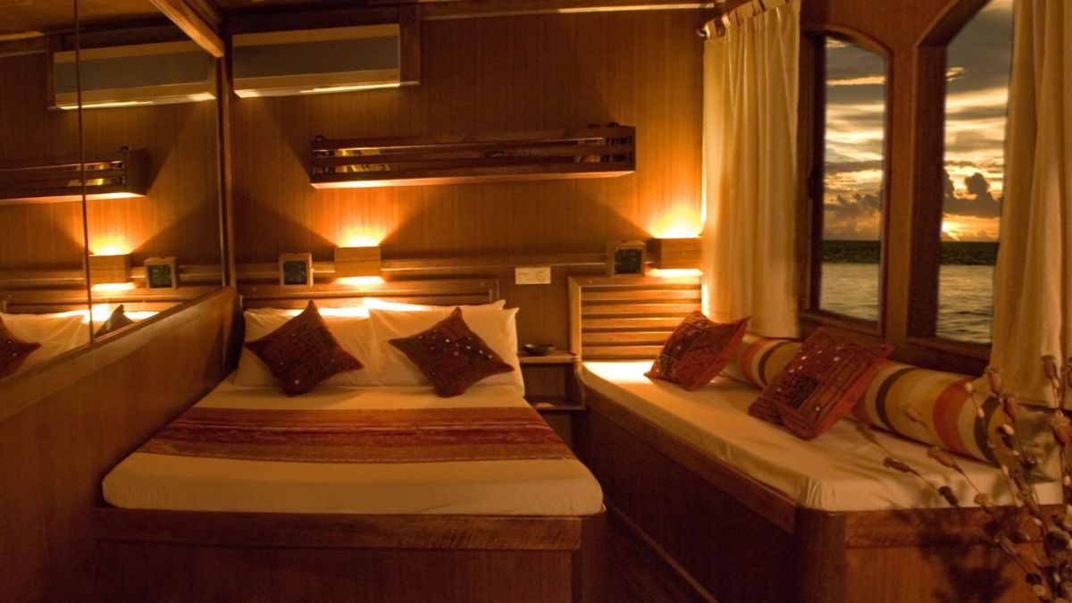 A cozy wooden cabin features a double bed with decorative cushions, soft lighting, and a comfortable seating area. Large windows offer a view of a sunset over the water, enhancing the warm atmosphere.