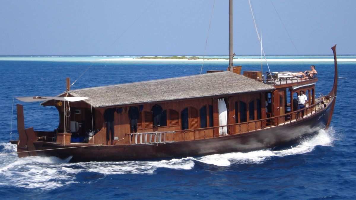 A wooden boat with a thatched roof sails over blue waters, surrounded by a tropical landscape. The boat features large windows and a deck with lounging areas, while a person stands on board.