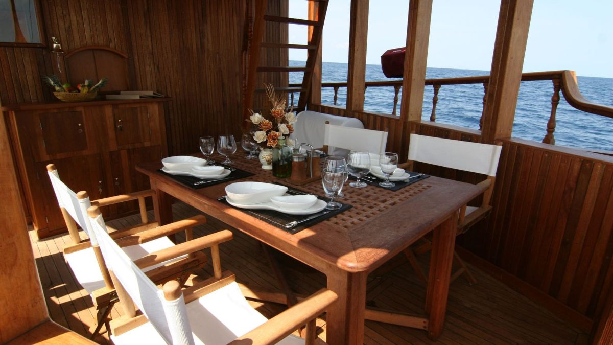 A wooden dining area on a boat featuring a large table set for a meal with white plates, glasses, and a floral centerpiece. The surrounding walls and furniture are made of polished wood, with a view of the sea visible through the windows.