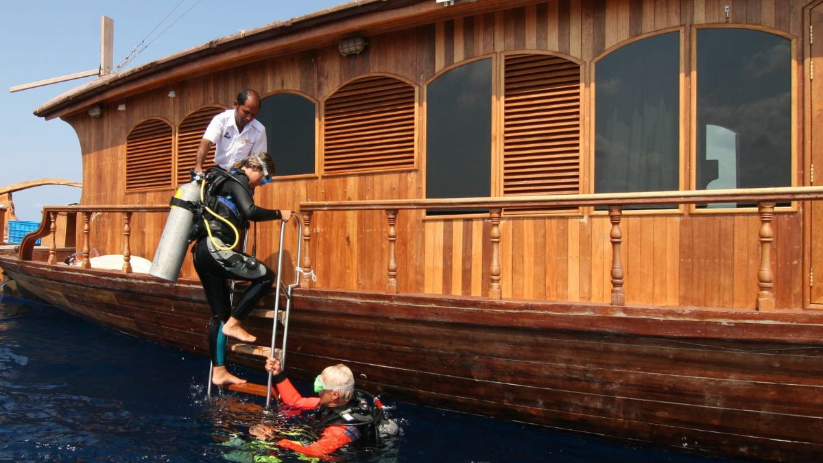 A diver in a wetsuit is being assisted out of the water by another diver while a crew member helps from a wooden boat with large windows and wooden accents. The scene takes place in a clear blue sea.