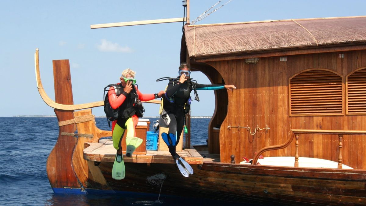 Two divers in wetsuits and fins are jumping off the side of a wooden boat into the ocean. The boat features a traditional design with a thatched roof. The sky is clear and blue.