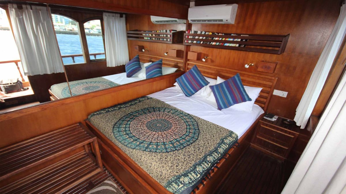 A cozy bedroom on a boat with wooden walls and furnishings. The bed is dressed with a patterned blanket and four decorative pillows. Two large windows let in natural light, revealing water views outside. A small bedside table and a bench are visible in the room.
