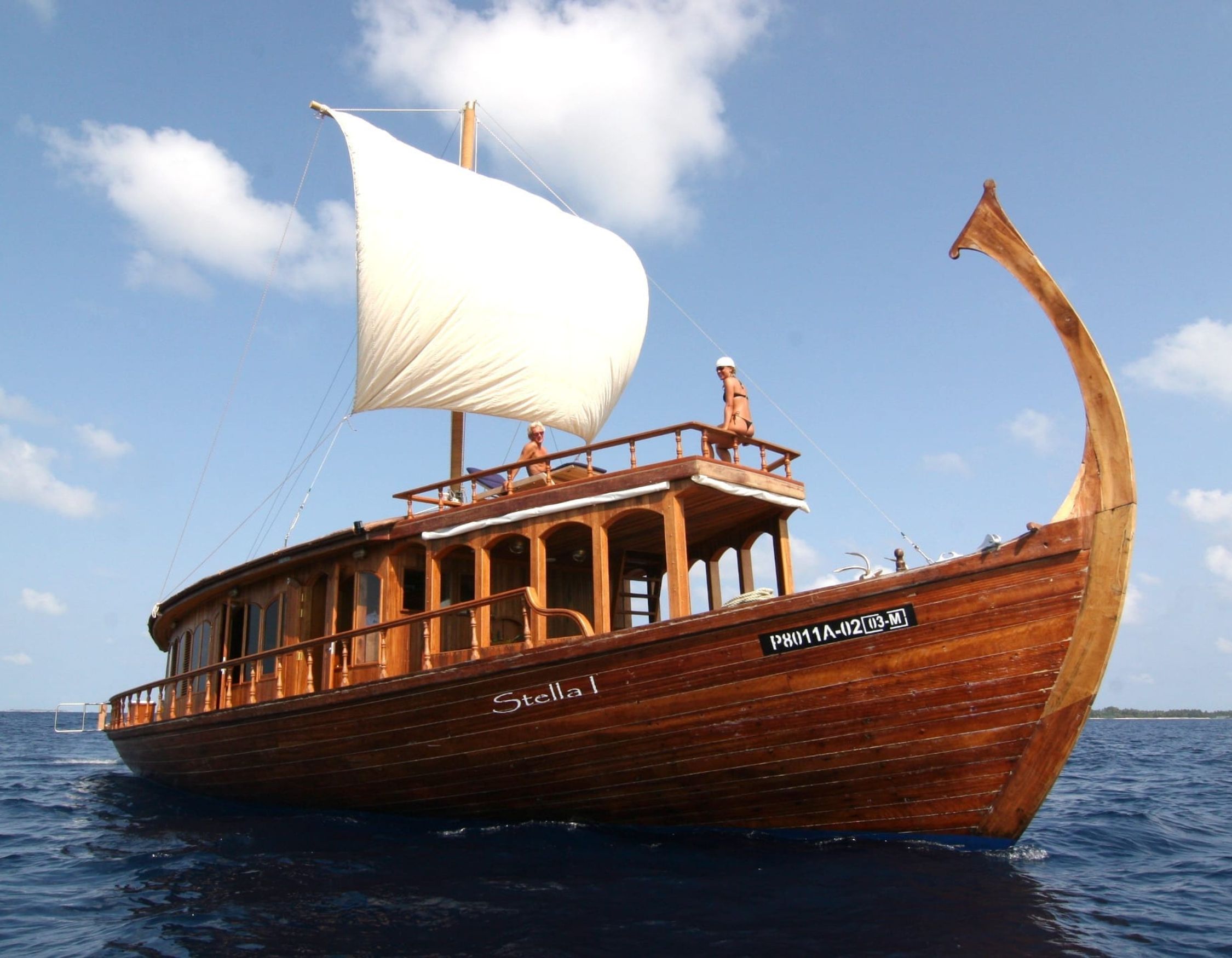 A wooden boat named "Stella I" sails on calm blue waters, featuring a large white sail and intricate carvings on its bow. Two individuals stand on deck, enjoying the scenic view under a partly cloudy sky.