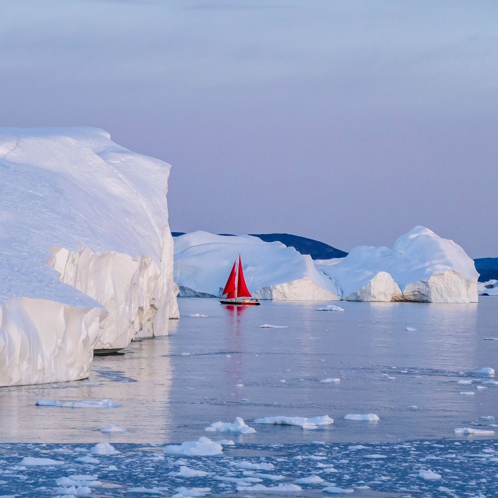A red sailboat navigates through icy waters surrounded by large white icebergs under a pale blue sky.
