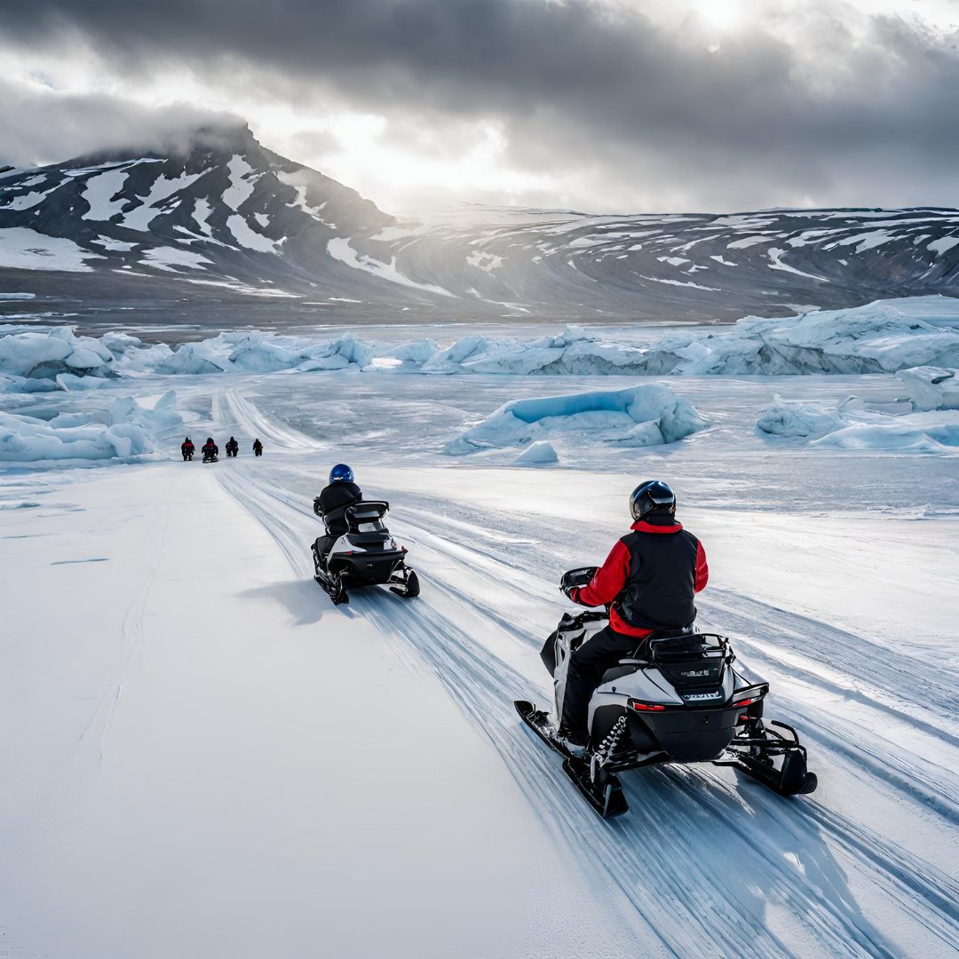 Snowmobiles traverse a snowy landscape, with riders dressed in winter gear. Frosty blue icebergs and dramatic mountain scenery are visible in the background, while dark clouds loom overhead.