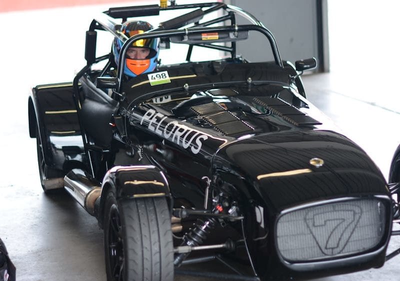 A driver in a racing helmet sits in a sleek, black race car with the number 7 displayed on the front. The car has an open-wheel design and is parked in a garage area, with other vehicles visible in the background.