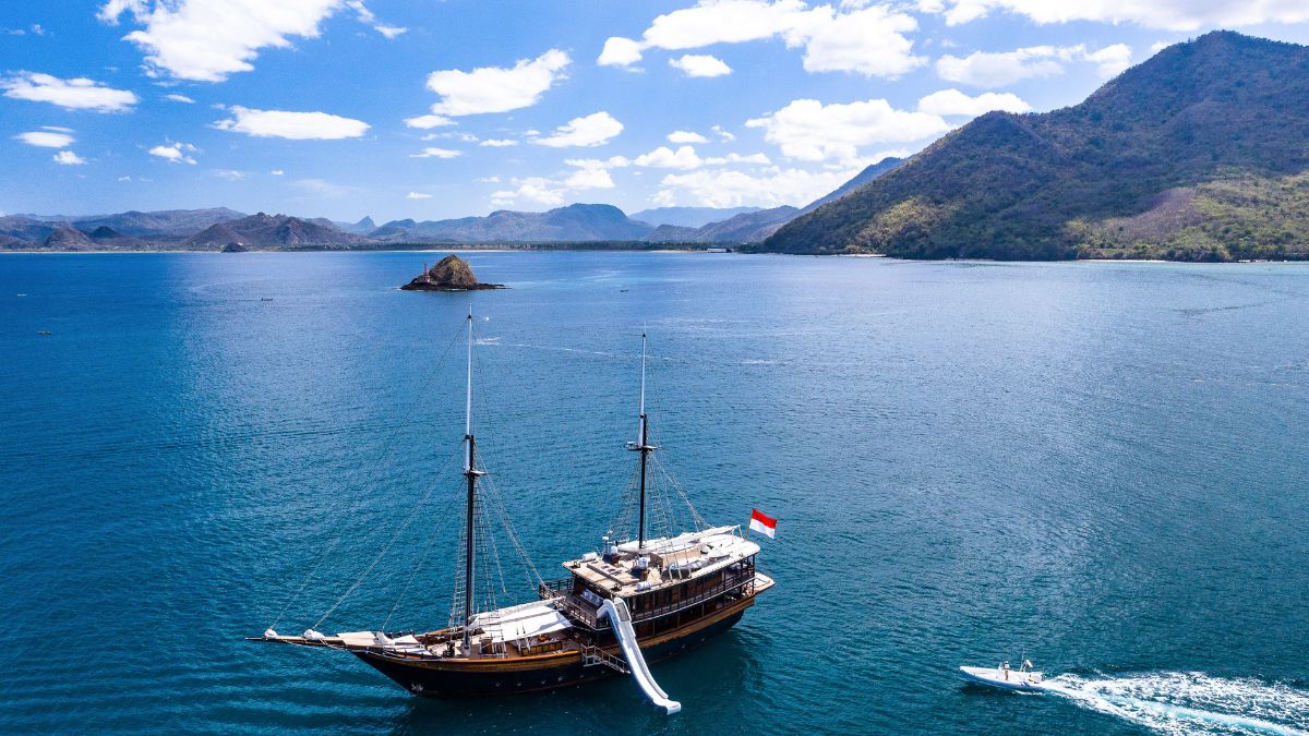 A traditional wooden ship sails through clear blue waters, with a small boat nearby. In the background, mountains rise under a partly cloudy sky.