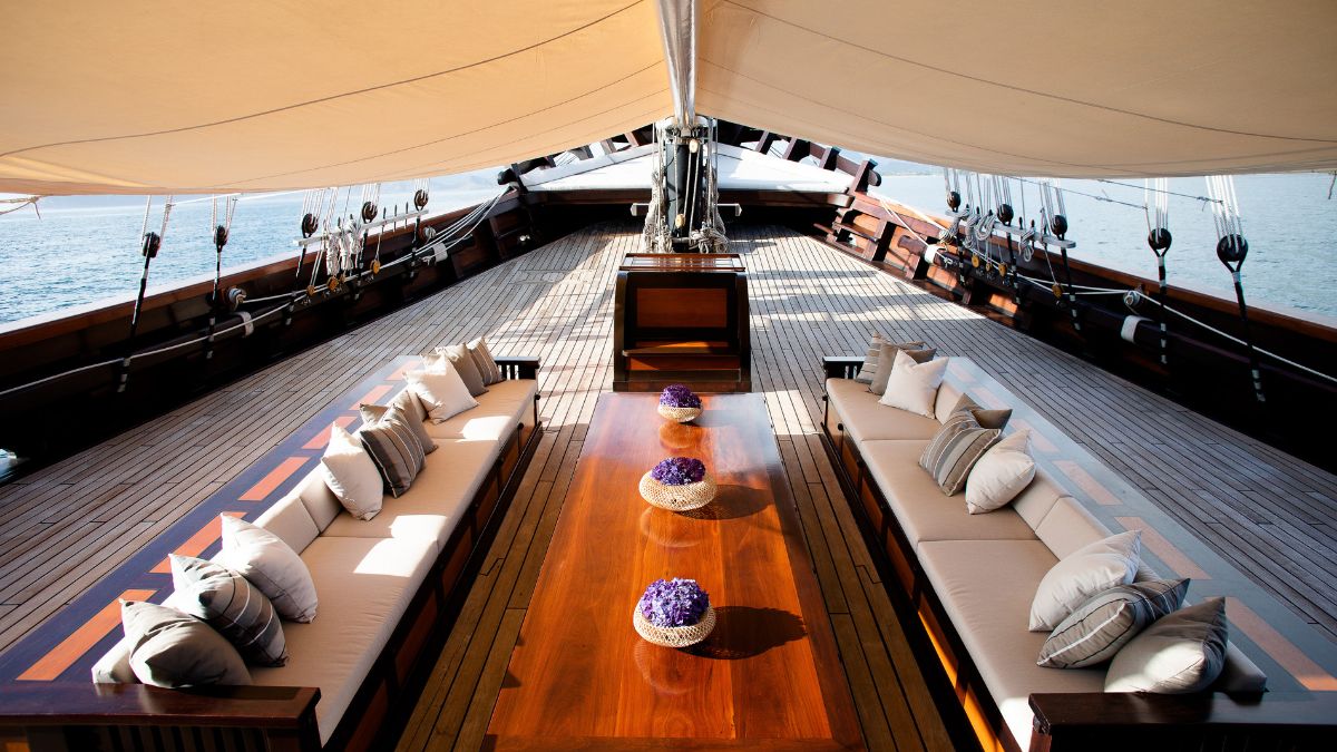 An elegant yacht deck featuring a spacious wooden table surrounded by comfortable seating. Brightly colored cushions enhance the relaxed atmosphere, with vases of flowers placed on the table. Large sails and the ocean are visible in the background.
