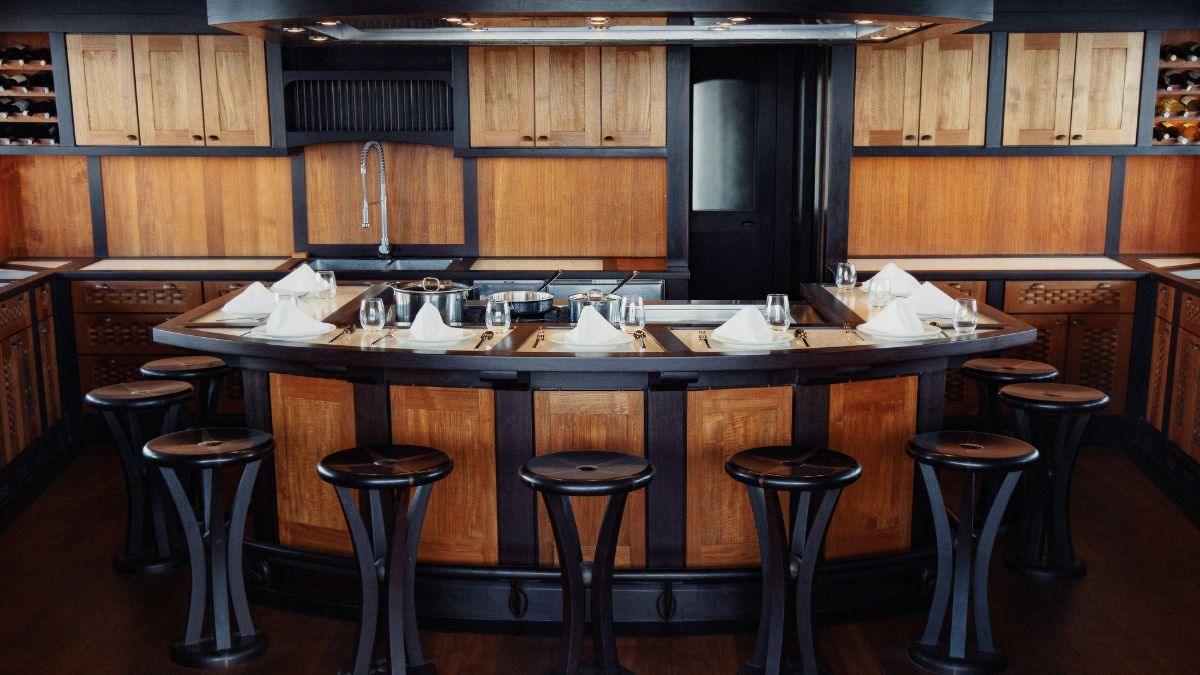 A modern kitchen featuring a central wooden island with cooking utensils and a sink, surrounded by high-backed stools. The cabinetry is dark with light wood accents, and there are neatly arranged napkins on the counter.