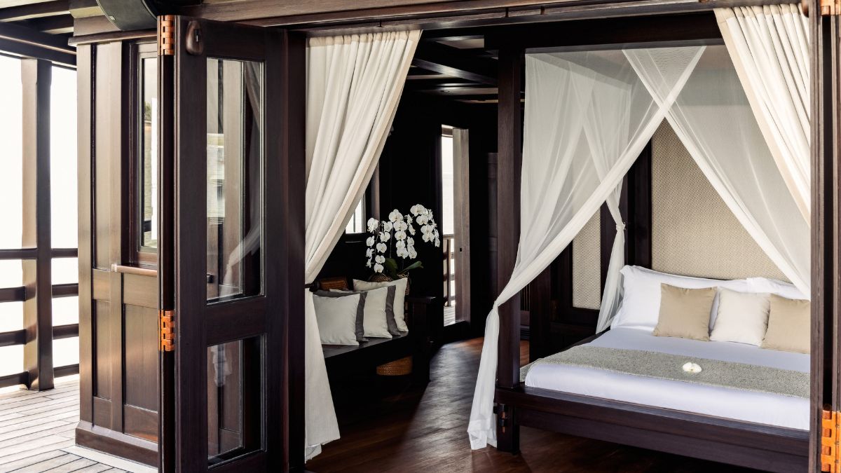 A luxurious bedroom featuring a four-poster bed with sheer curtains, elegantly decorated with neutral-toned cushions. The space includes a cozy sitting area with a flower arrangement, all set within a stylish wooden interior and large windows that allow natural light.