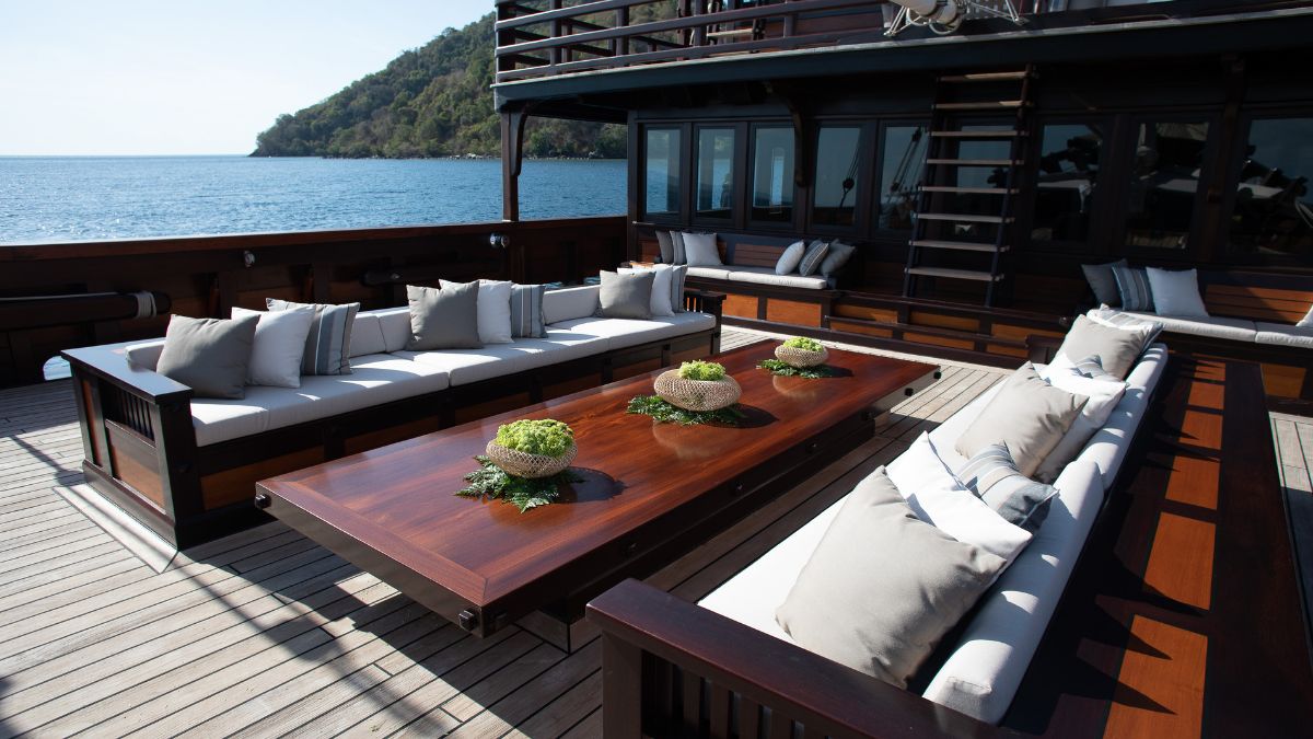 Luxurious outdoor lounge area on a yacht, featuring plush seating with grey cushions around a large wooden table, set against a serene sea view.
