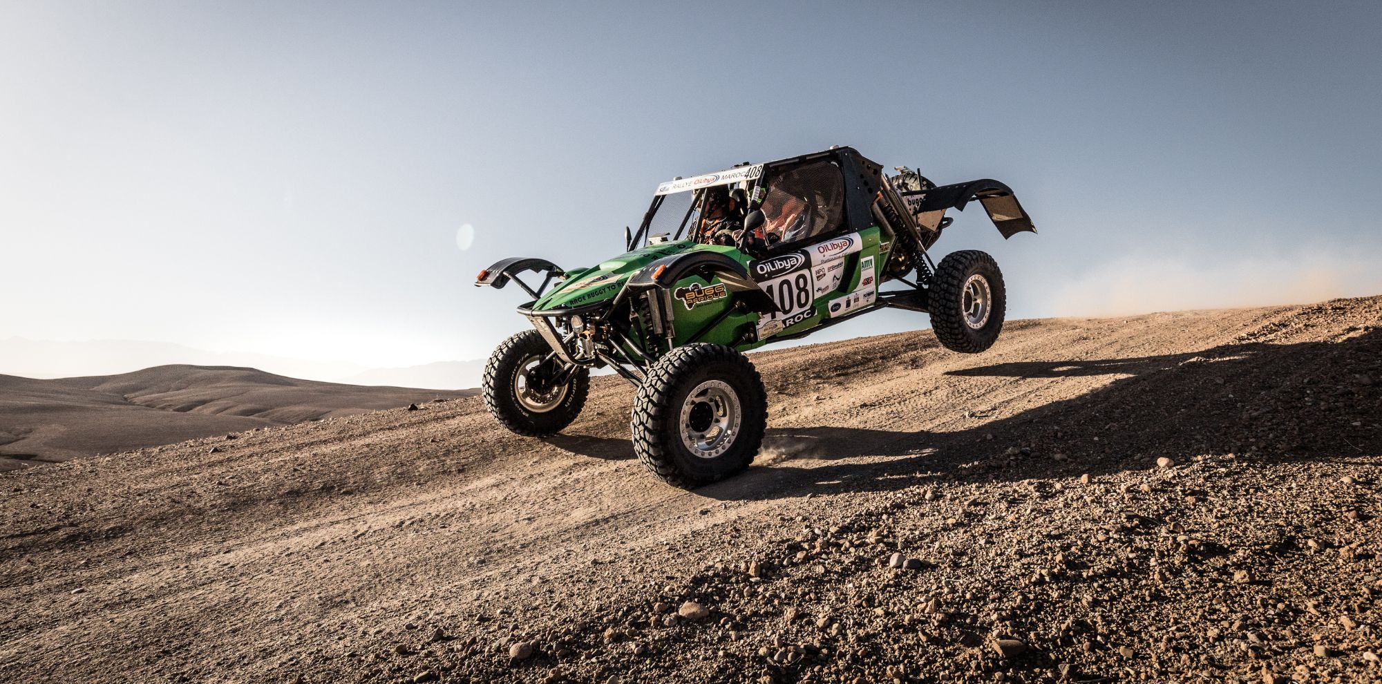 A green off-road vehicle jumps over a dirt path in a desert landscape, dust rising behind it under a clear blue sky.
