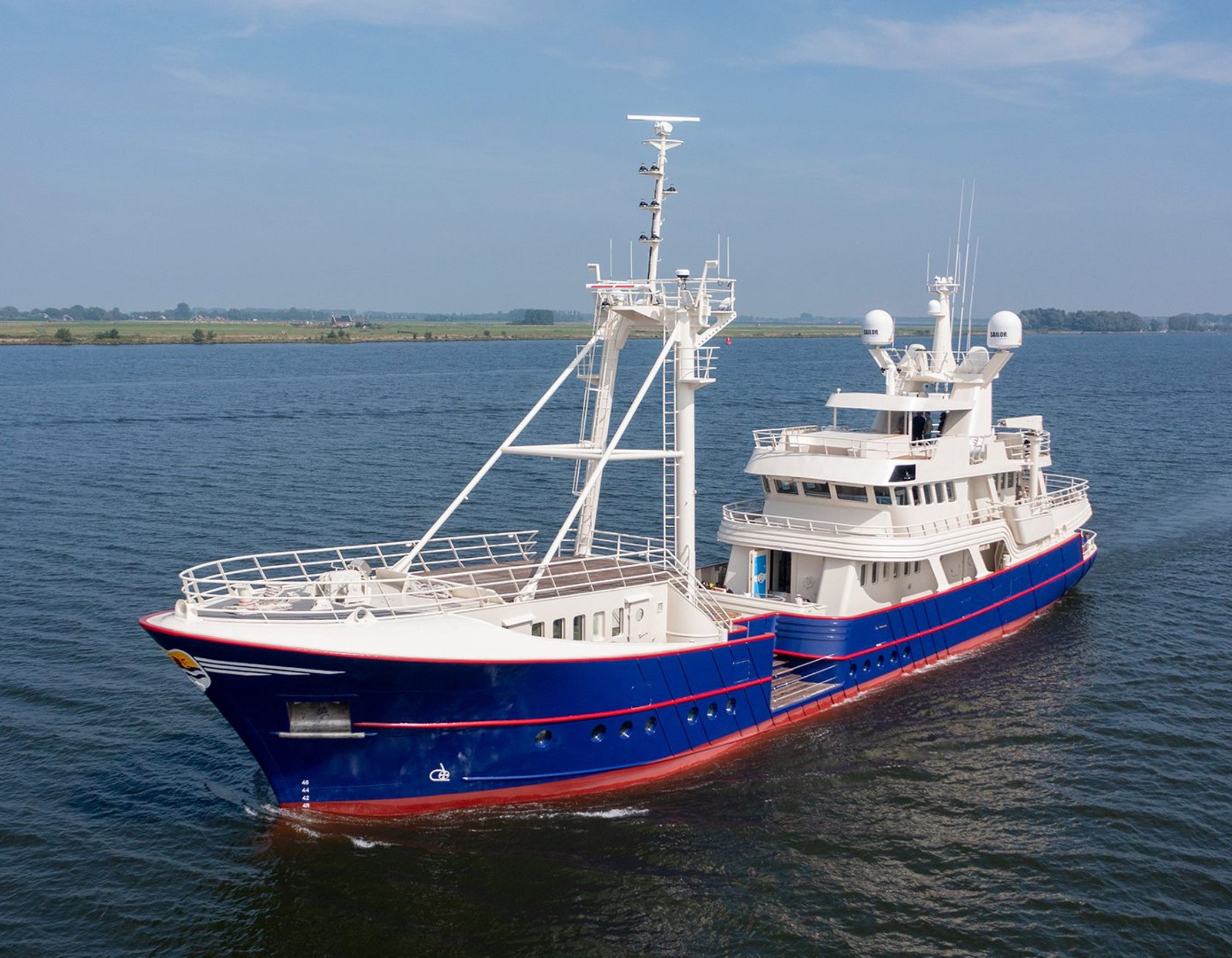 A large motor yacht with a sleek blue and white hull cruising on calm waters. The vessel features multiple decks, antennas, and radar equipment, set against a clear sky and distant green landscape.