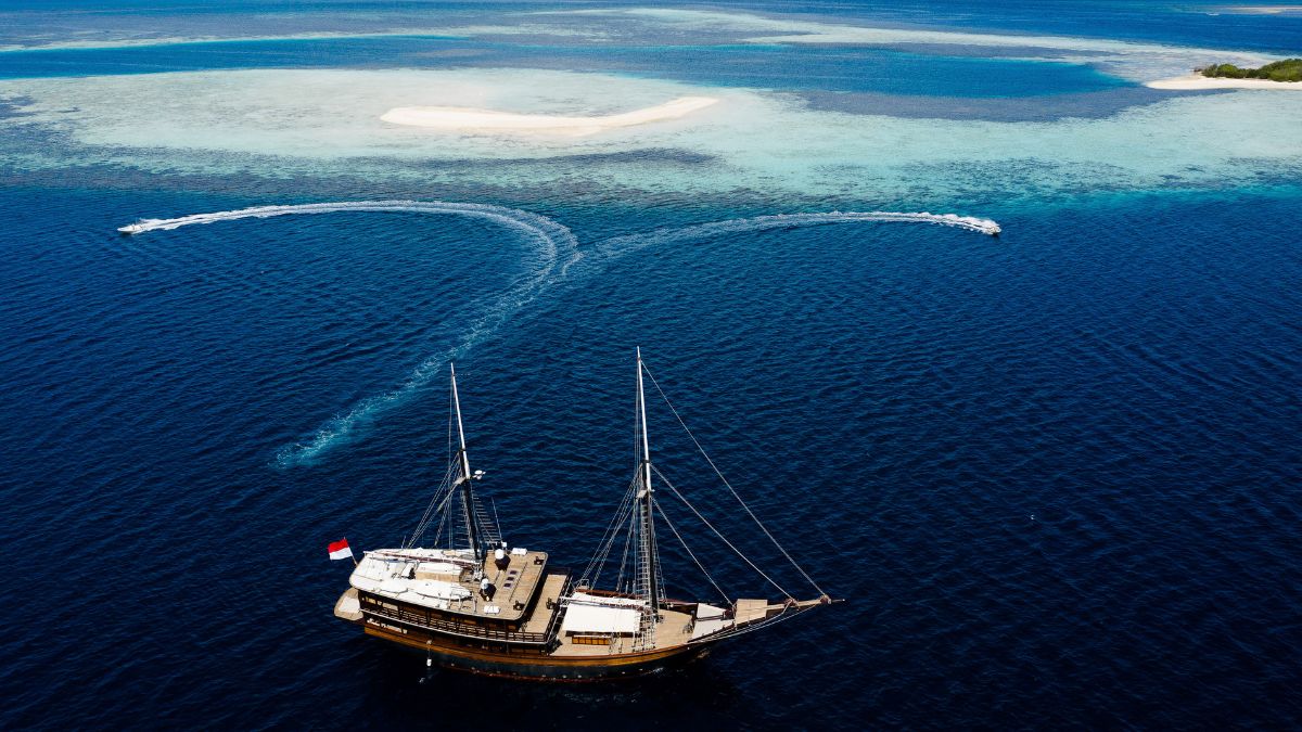 A large wooden sailing ship with a flag sails through deep blue waters near a sandy island, with two speedboats creating white trails in the water.