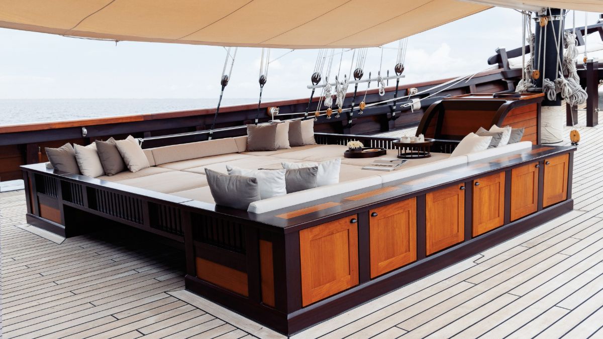 A spacious wooden deck on a yacht features a large, comfortable seating area with soft cushions and a low table. Nautical elements, such as ropes and sails, are visible in the background against an ocean backdrop.