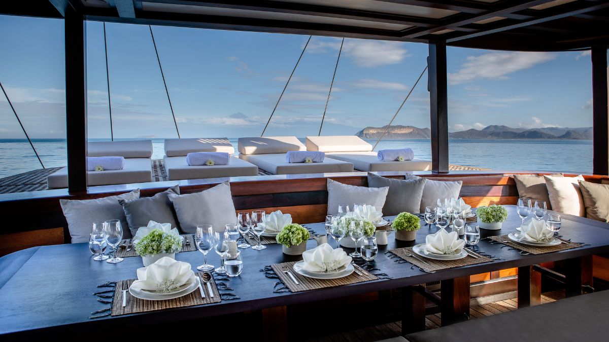 A beautifully set dining table on a boat, featuring elegant tableware, flower arrangements, and glassware. In the background, serene water and mountains are visible under a blue sky.
