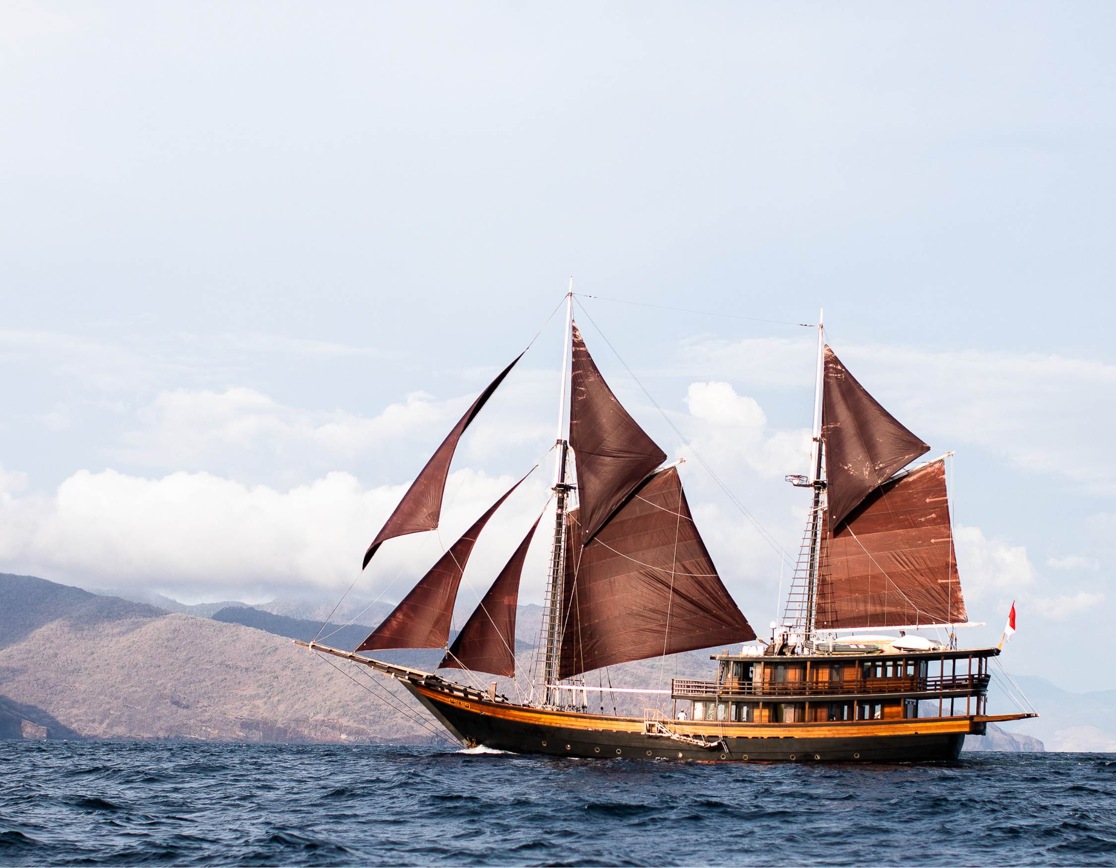 A majestic sailing ship with large brown sails glides across calm waters, set against a backdrop of distant mountains and a blue sky.