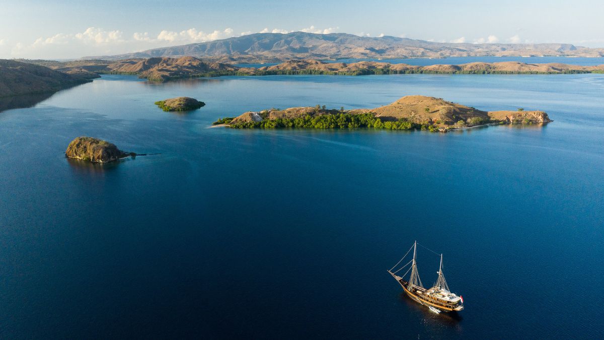 A lone sailing ship anchored near a rocky coast, surrounded by calm blue waters and lush green islands, with mountains in the background under a clear sky.