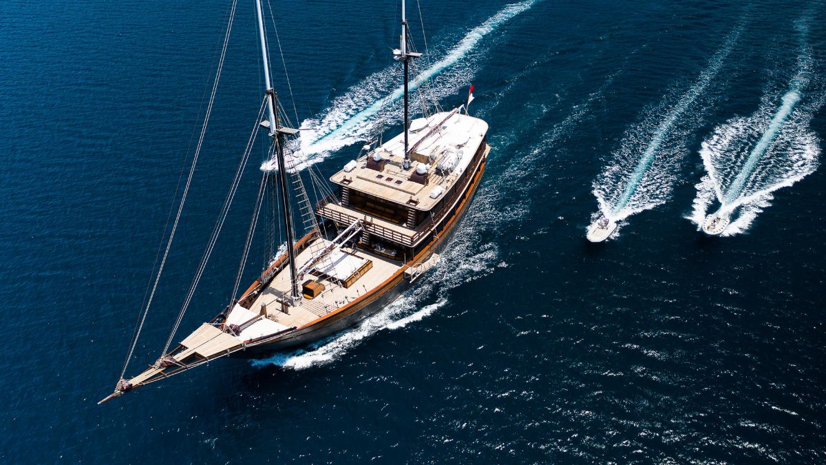 A large sailing yacht glides through deep blue waters, leaving a wake behind. Two smaller motorboats trail closely behind, creating additional waves.