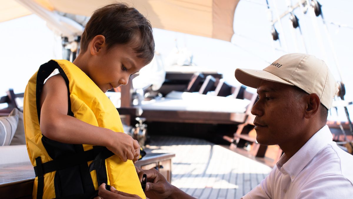 A young boy is wearing a yellow life jacket while a man helps him fasten it. They are on a boat with a sail in the background.