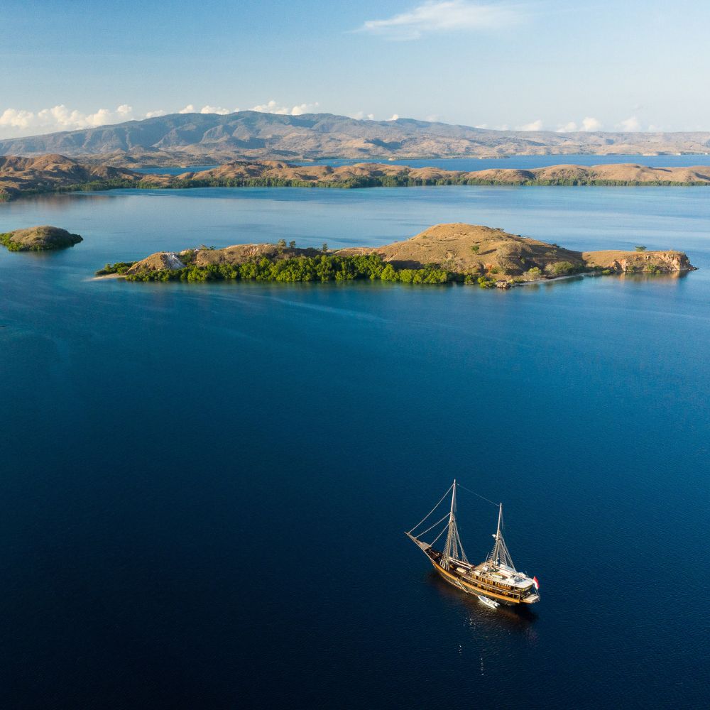 A classic sailing ship glides through calm blue waters, surrounded by lush green islands and distant mountains under a clear sky.