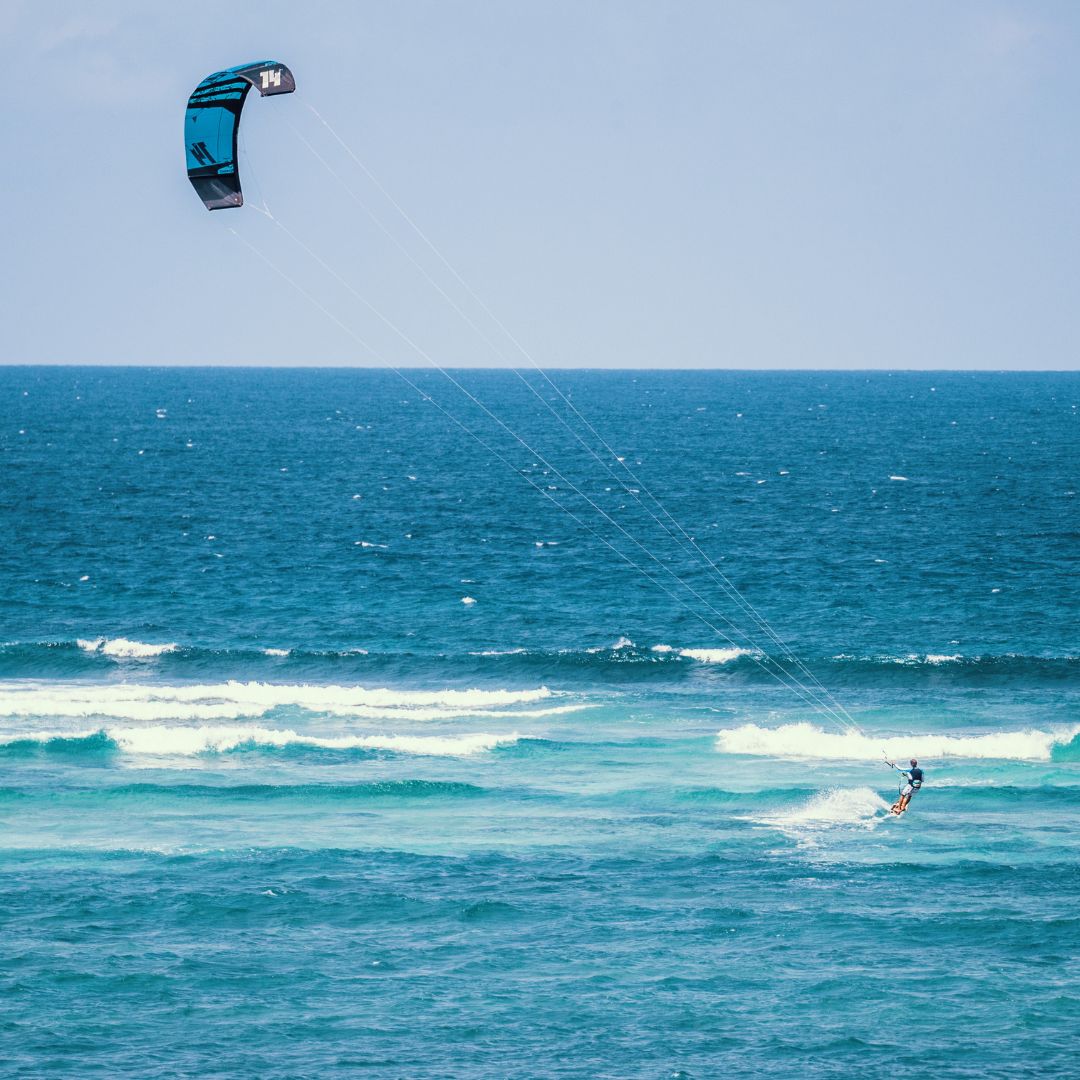 A kitesurfer performs tricks on a vibrant blue ocean, with waves gently rolling in beneath a sunny sky and a large, colorful kite soaring above.