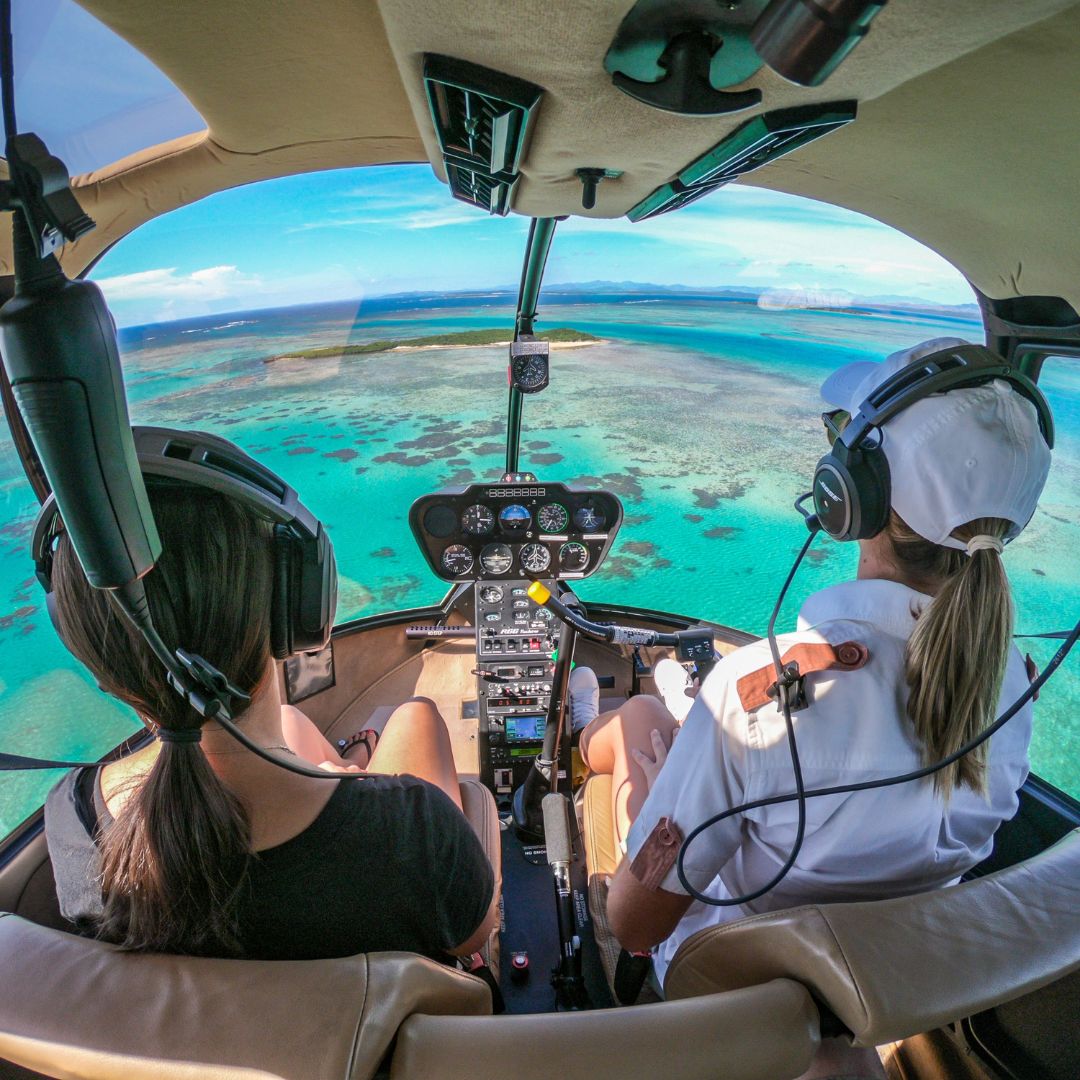 Two people wearing headsets sit in the cockpit of a helicopter, overlooking vibrant turquoise waters and coral reefs below. The landscape features islands and a clear blue sky in the background.