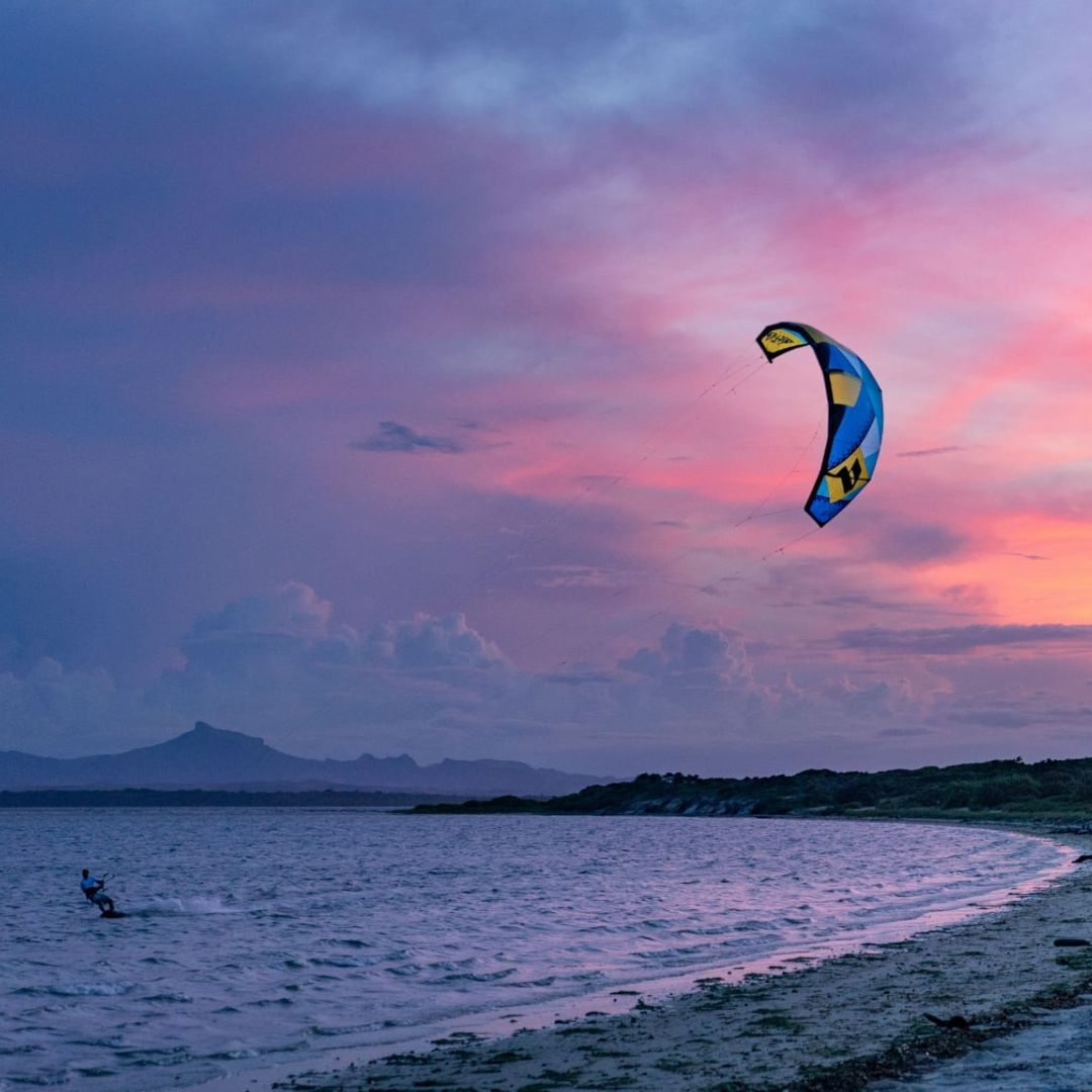 A kite surfer rides the water at sunset, with a colorful kite soaring above against a backdrop of pink and purple skies. Mountains silhouette in the distance.