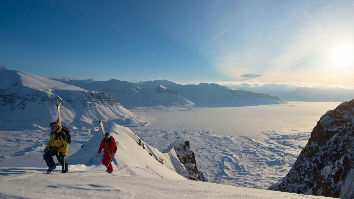 Two climbers ascend a snowy slope in a mountainous landscape. The scene includes vast glacial expanses and distant peaks under a bright blue sky with wispy clouds. Sunlight casts a warm glow over the icy terrain.