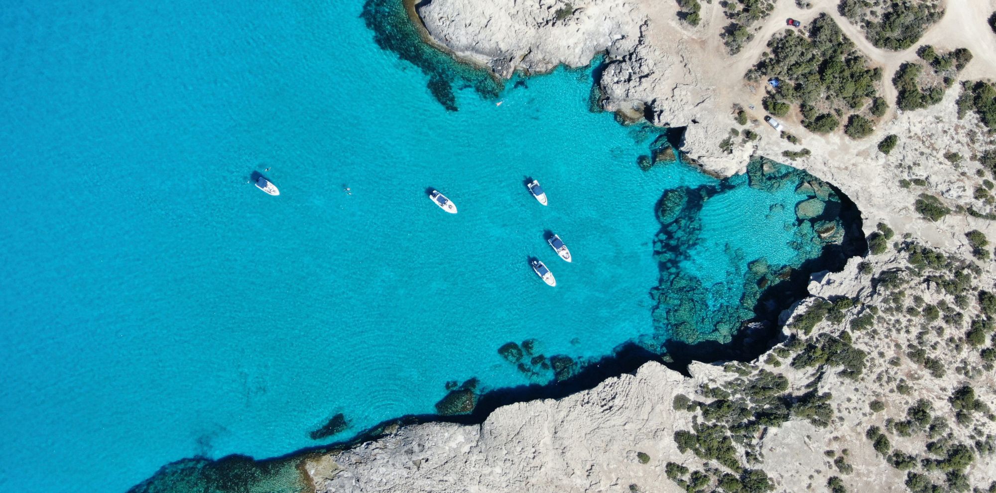 Aerial view of a crystal-clear turquoise sea with several boats anchored near rocky shores. The coastline features a mix of sand and rocky terrain.