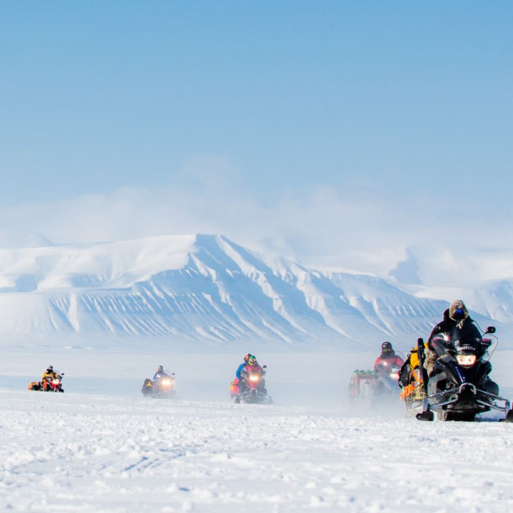 A group of snowmobiles traversing a snowy landscape with majestic mountains in the background under a clear blue sky. Snow is being kicked up by the vehicles, creating a sense of adventure.