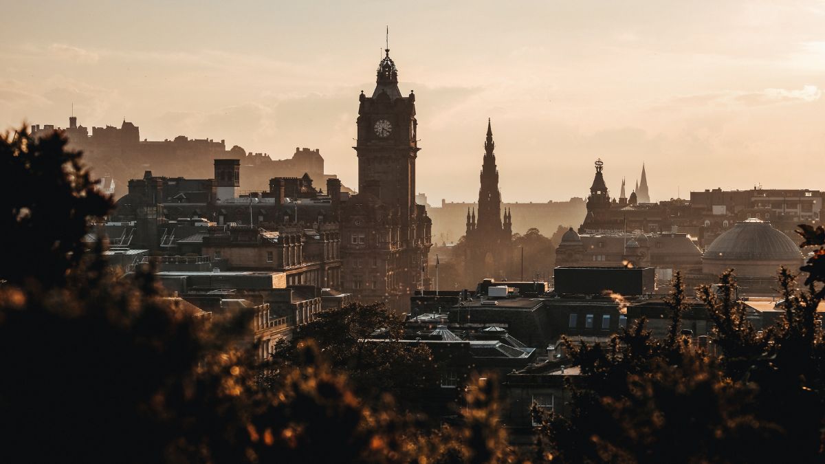 A panoramic view of Edinburgh's skyline at sunset, featuring the iconic Balmoral Clock Tower and various historical buildings silhouetted against a golden sky. The landscape is framed by foliage in the foreground.