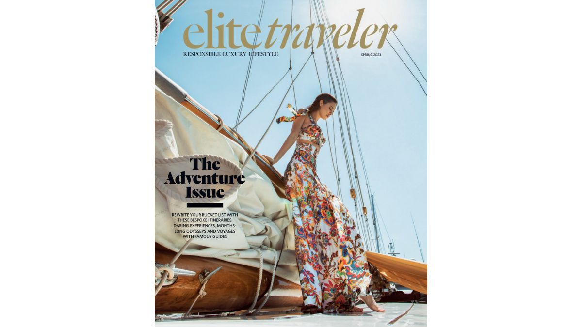 A woman in a vibrant, floral maxi dress poses elegantly on a yacht, set against a bright blue sky and white sail. The magazine cover states "elite traveler" and features the title "The Adventure Issue."