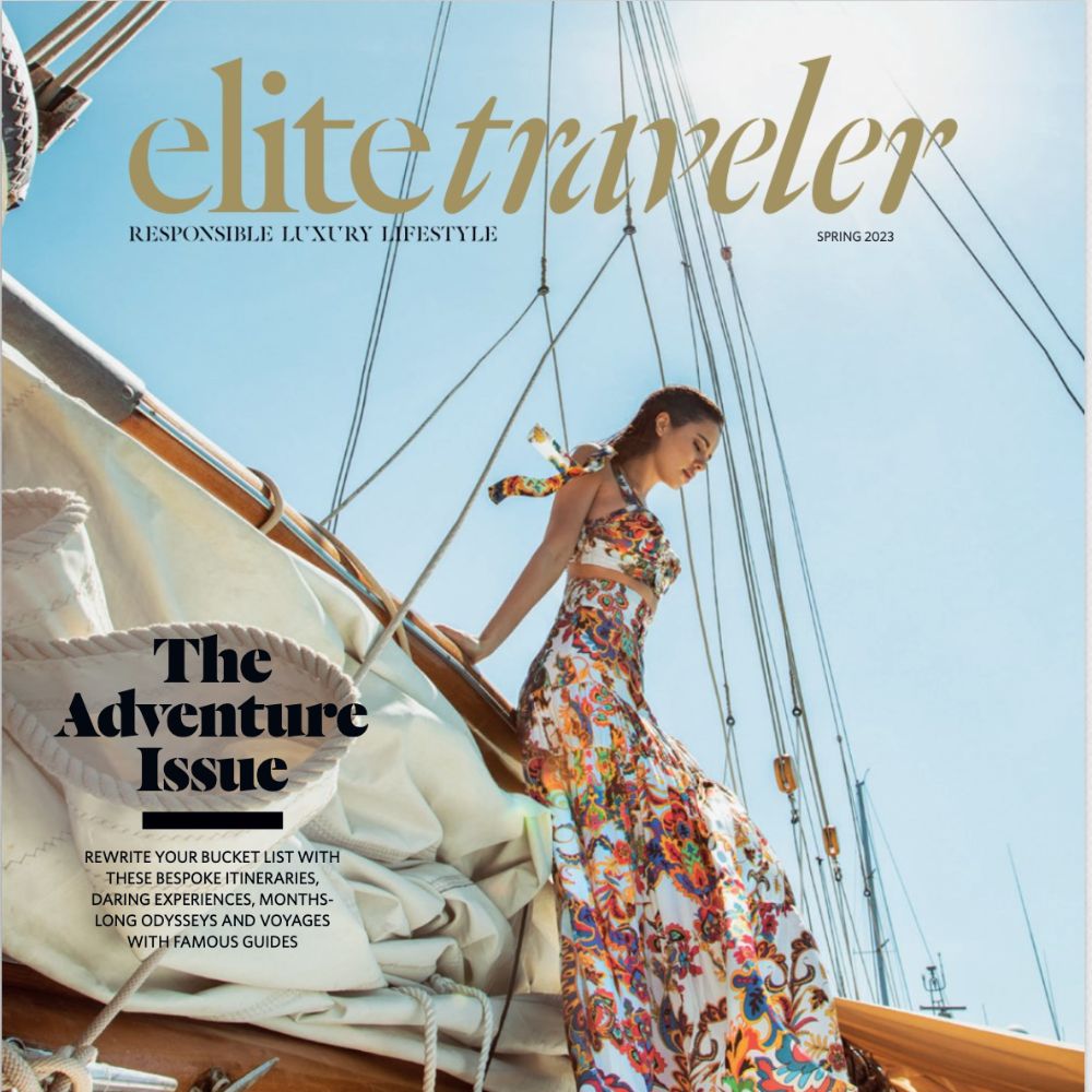 Woman in a colorful, flowing dress leans against the mast of a sailboat, with the magazine title "Elite Traveler" and headlines for the Spring 2023 issue prominently displayed. The theme "The Adventure Issue" suggests luxury travel and adventure experiences.