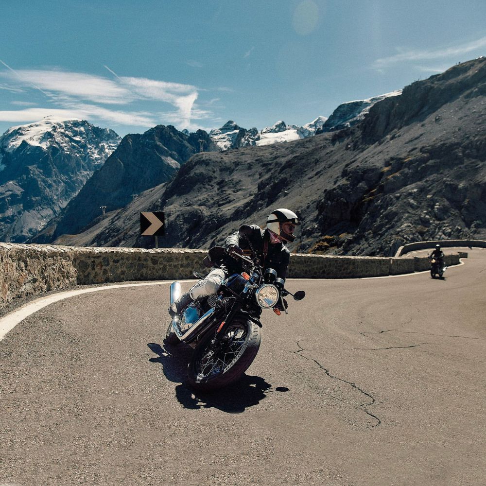 A motorcyclist leans into a curve on a winding mountain road, with snow-capped peaks and rocky cliffs in the background under a clear blue sky. Another motorcycle follows in the distance.
