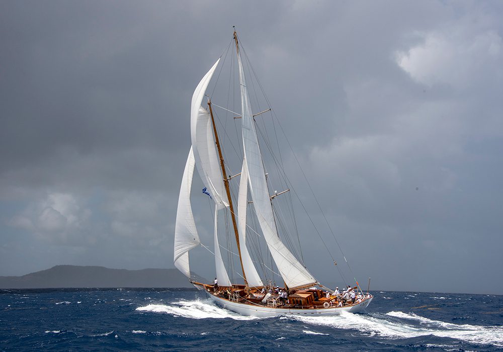 A classic sailboat with large white sails cuts through choppy blue waters under a cloudy sky. Waves splash against the hull as the boat leans to one side, with passengers visible on deck.