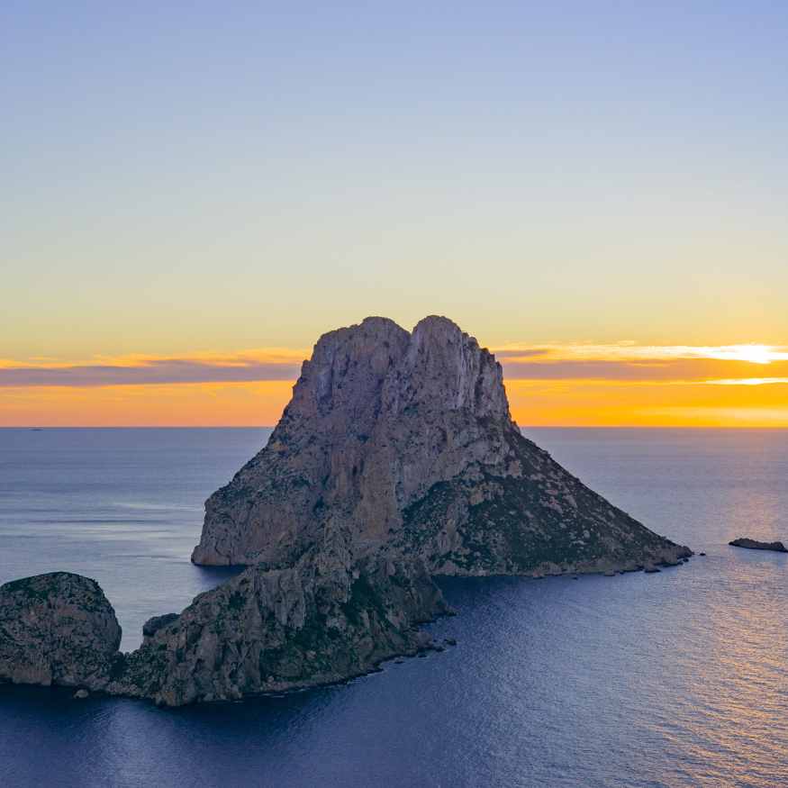 A prominent rocky island rises from the ocean, surrounded by calm waters at sunset. The sky is painted with soft hues of orange and purple, casting a serene glow over the landscape.