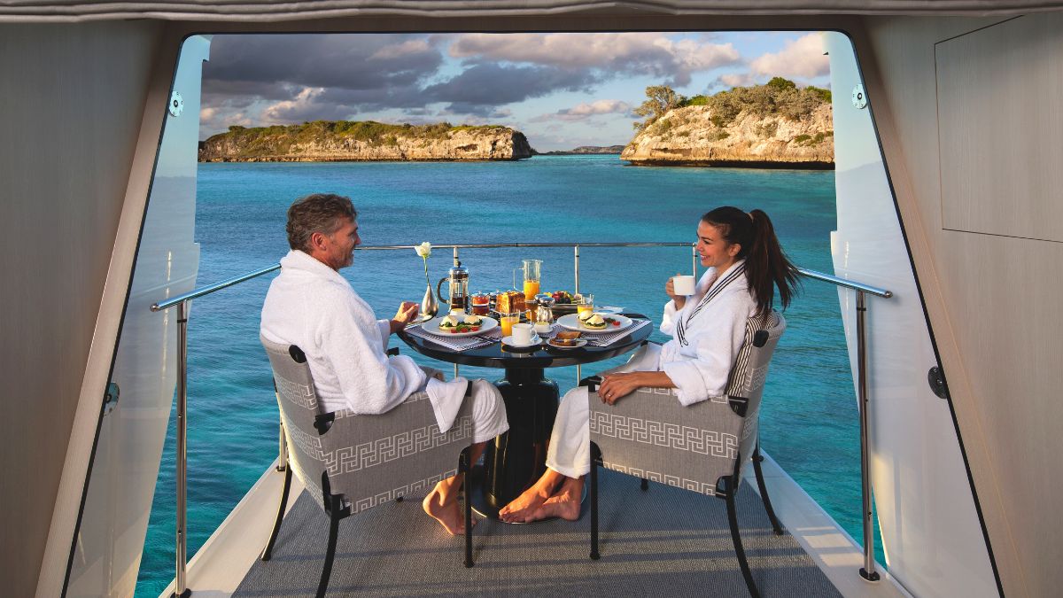 A couple enjoys a luxurious breakfast on a yacht balcony overlooking a serene turquoise sea and rocky islands. They sit at a table set with food and drinks, both dressed in white robes, relaxed and smiling.