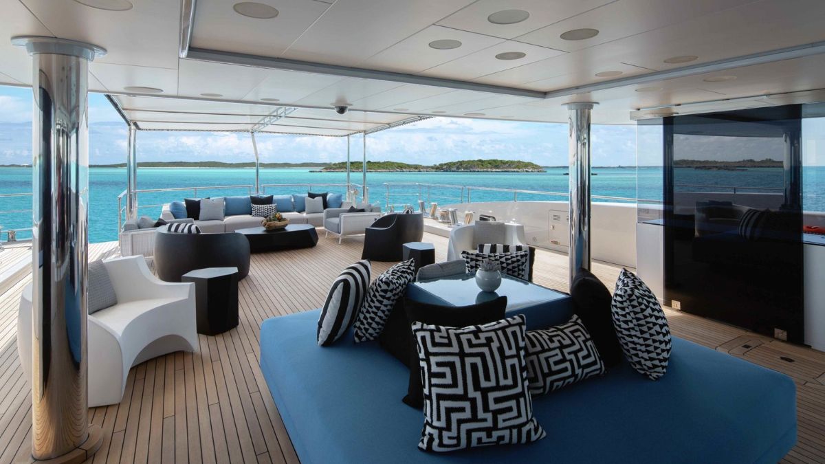 A stylish outdoor lounge area on a yacht features modern seating with patterned cushions, a coffee table, and panoramic views of the turquoise sea and distant islands. The deck is made of wooden planks, and the space is bathed in natural light.