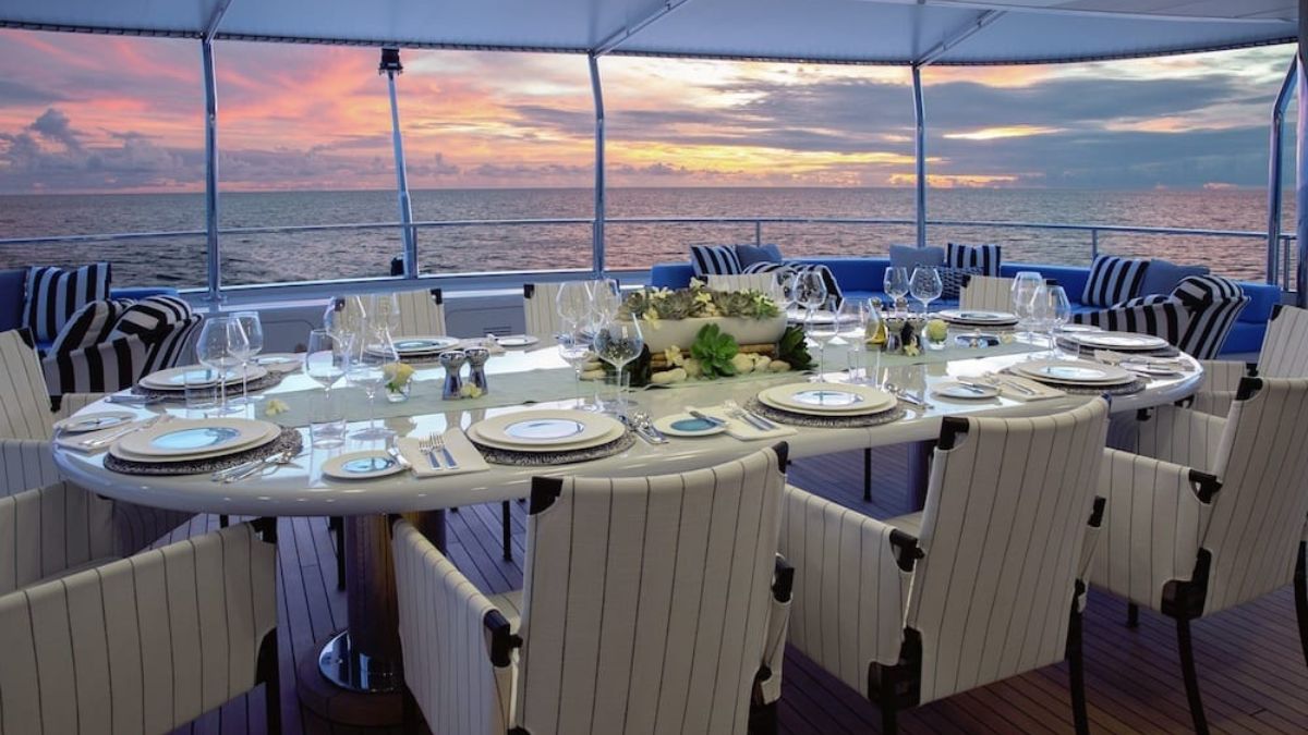 A beautifully set dining table with elegant tableware overlooks a serene ocean view during sunset. The table is surrounded by striped chairs, and the sky is painted with vibrant colors as the sun sets on the horizon.