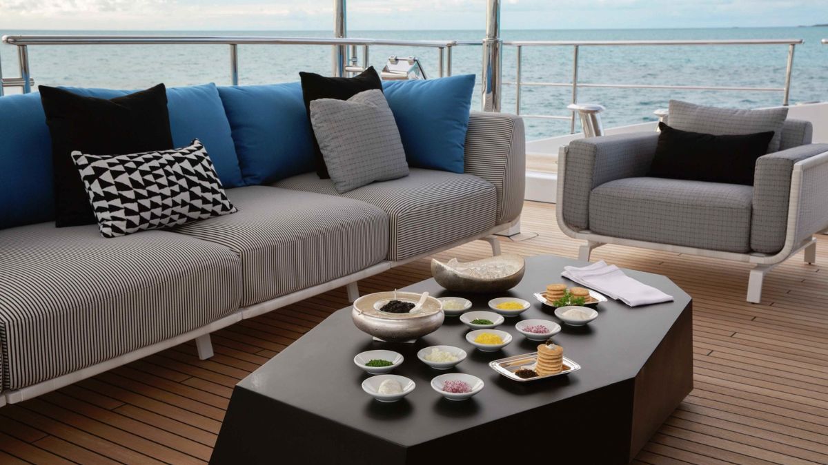 A stylish outdoor seating area on a yacht, featuring a modern couch with black and white striped cushions and blue throw pillows. A sleek black coffee table is adorned with an array of condiments and snacks, set against a serene ocean backdrop.