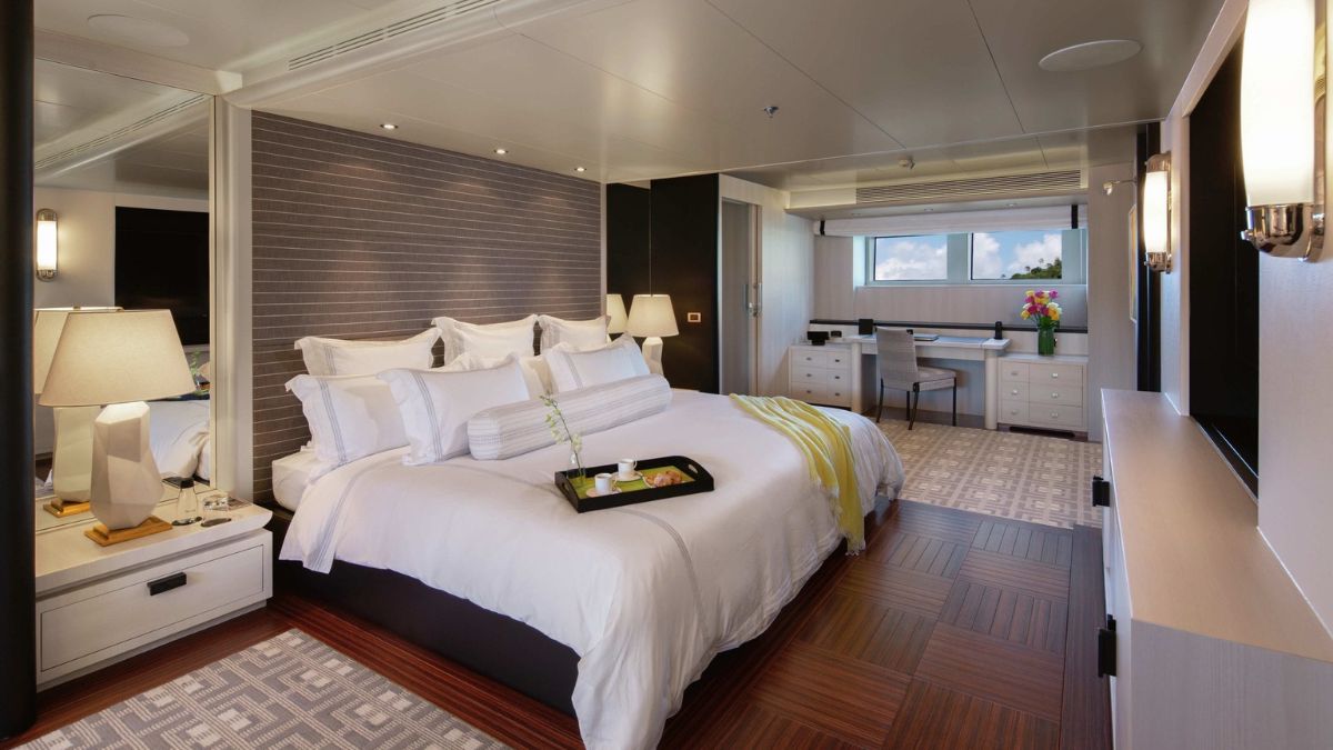 A modern bedroom featuring a large bed with white linens, a decorative yellow throw, and a tray with snacks on the bedside table. The room has a stylish design with dark wood accents, a window allowing natural light, a work desk, and flowery decor.