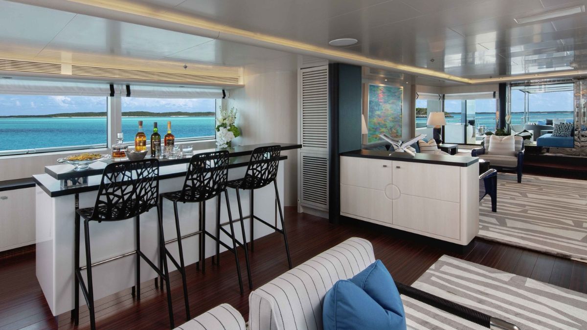 A modern yacht interior featuring a sleek bar with three high stools, a countertop with bottles, and glasses. Large windows offer views of turquoise waters, while cozy seating areas with striped patterns provide a relaxed atmosphere.